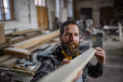 Ein reifer männlicher Schreiner trägt ein Holzbrett in einer Schreinerwerkstatt, ein kleines Geschäftskonzept. - HPIF06405
