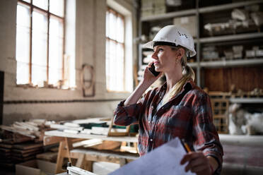 A female engineer holding blueprints and making phone call indoors in carpentry workshop. - HPIF06419