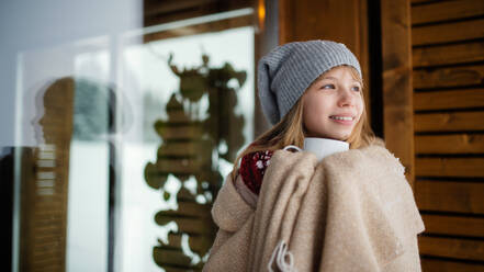 Porträt eines jungen Mädchens in eine Decke eingewickelt mit einem Getränk im Freien vor einem Holzhaus in der winterlichen Natur. - HPIF06631