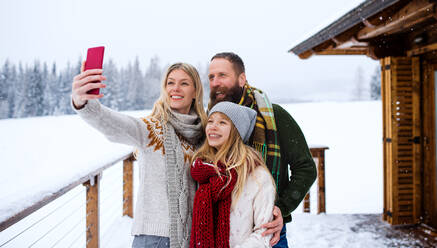 Eine fröhliche Familie hält eine Wintererinnerung fest, während sie ein Selfie vor ihrer Berghütte mit ihrer kleinen Tochter macht - HPIF06632
