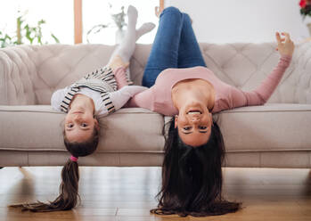 Portrait of mother with small daughter on sofa indoors at home, looking at camera upside down. - HPIF06674