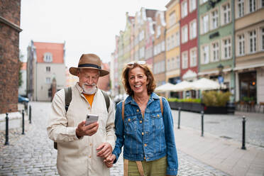 A portrait of happy senior couple tourists smiling, holding hands, using smartphone outdoors in historic town - HPIF06701
