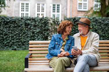Ein glückliches, verliebtes älteres Paar sitzt auf einer Bank und benutzt sein Smartphone im Freien in der Stadt, Kopierraum. - HPIF06731