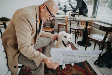 Ein glücklicher älterer Mann sitzt in der Cafeteria und liest mit seinem Hund Zeitung. - HPIF06846