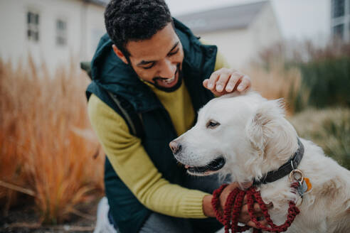 Happy young man stroking his dog outdoors in city park, during a cold autumn day. - HPIF06848