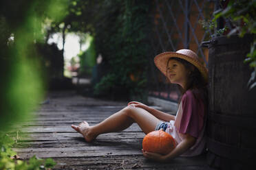 Ein kleines Mädchen mit Hut sitzt und hält einen Bio-Kürbis im Freien auf einem Bauernhof. - HPIF06911