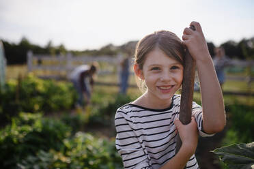 Porträt eines glücklichen kleinen Bauernmädchens mit Gartengerät im Freien auf einem Gemeinschaftsbauernhof, das in die Kamera schaut. - HPIF06930