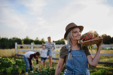 Eine ältere Bäuerin trägt einen Korb mit selbst angebautem Gemüse im Freien auf einer Gemeinschaftsfarm. - HPIF06956