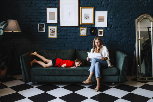 Woman with documents sitting by boy lying on sofa at studio - MDOF00674