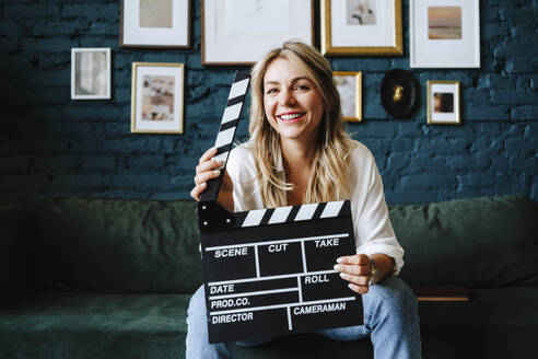 Happy actress holding clapboard sitting on sofa at film set - MDOF00697