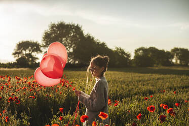 Mädchen hält Luftballons inmitten von Mohnblumen bei Sonnenuntergang - ALKF00143