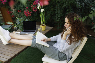 Frau, die im Garten ein Buch liest und mit ihrem Smartphone telefoniert - TYF00761