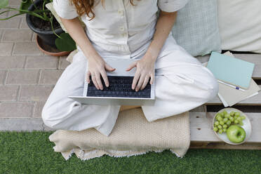 Freelancer working on laptop sitting cross-legged on crate at backyard - TYF00765