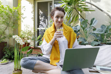 Smiling freelancer with laptop drinking water in backyard - TYF00777