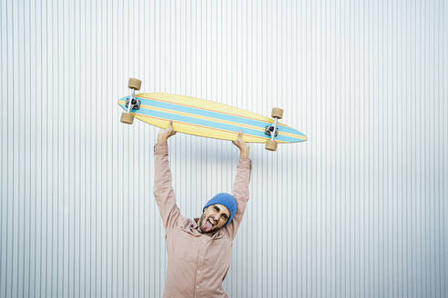 Man with tongue out holding longboard in front of white wall - RCPF01691