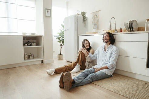 Happy couple sitting on floor in kitchen at home - RCPF01715