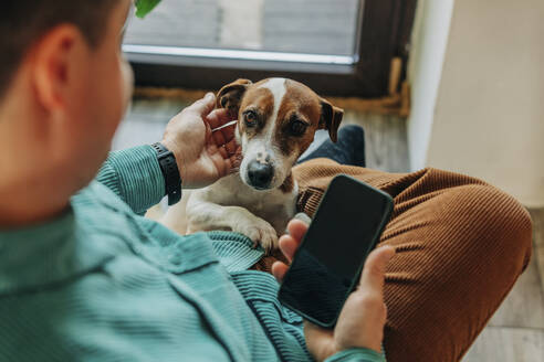 Mann hält Smartphone und streichelt Hund zu Hause - VSNF00556