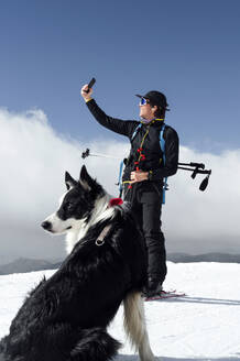 Man taking selfie with smart phone with dog sitting on snow - PGF01473