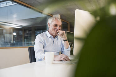 Smiling senior businessman with hand on chin working on computer at office - UUF28255