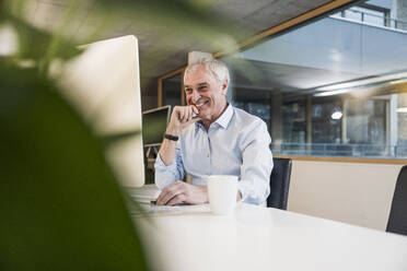 Lächelnder älterer Geschäftsmann mit Hand am Kinn bei der Arbeit am Computer im Büro - UUF28256