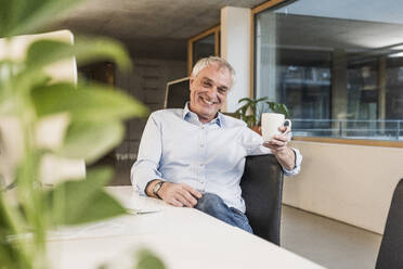 Happy senior businessman with mug sitting at desk in office - UUF28258