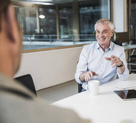 Happy senior businessman talking to colleague sitting at desk in office - UUF28267