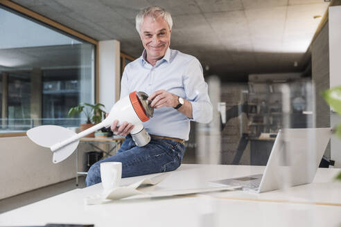 Smiling senior engineer holding wind turbine rotor sitting on desk at office - UUF28275