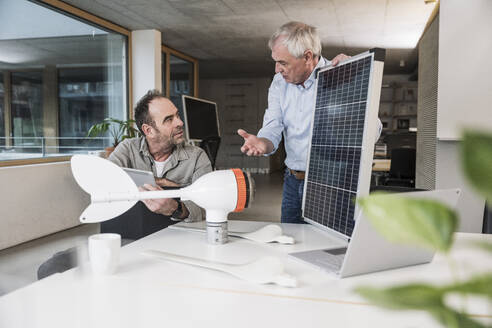 Geschäftsmann an einem Sonnenkollektor, der mit einem Kollegen über einen Windradrotor im Büro diskutiert - UUF28282
