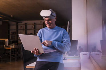 Lächelnder Geschäftsmann, der ein Virtual-Reality-Headset trägt und einen Laptop im Büro benutzt - UUF28306