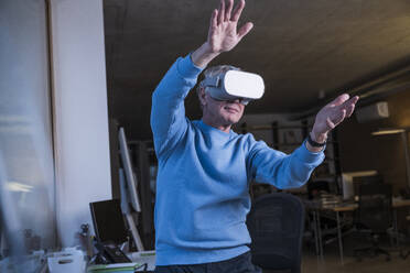 Elderly businessman wearing virtual reality headset and gesturing at office - UUF28310