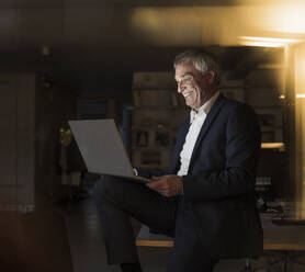 Happy businessman with laptop sitting at office - UUF28321