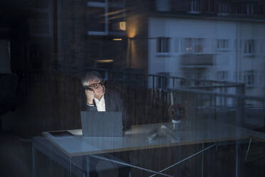 Businessman sitting with laptop at desk in office seen through window - UUF28331