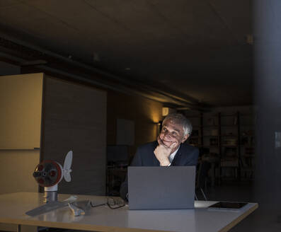 Smiling businessman with hand on chin sitting at desk in office - UUF28333