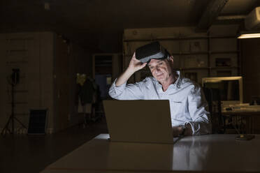 Senior businessman holding virtual reality headset working on laptop at office - UUF28345