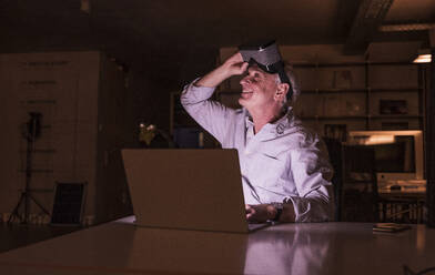 Lächelnder Geschäftsmann, der ein Virtual-Reality-Headset hält und auf sein Büro schaut - UUF28346