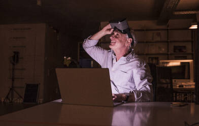 Smiling businessman holding virtual reality headset looking up at office - UUF28346