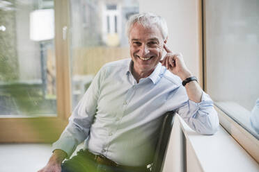 Happy businessman sitting on chair near window at office - UUF28349