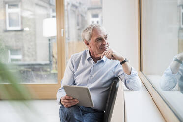 Nachdenklicher älterer Geschäftsmann mit Tablet-PC am Fenster sitzend im Büro - UUF28352