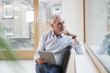 Thoughtful senior businessman with tablet PC sitting near window at office - UUF28352