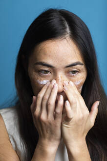 Woman with freckles applying lotion on face against blue background - MRAF00924