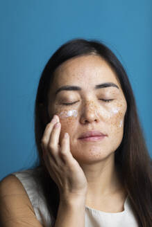 Woman with eyes closed applying moisturizer against blue background - MRAF00925