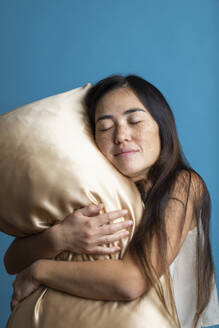 Woman with eyes closed hugging golden pillow against blue background - MRAF00926