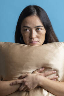 Thoughtful woman resting chin on silk pillow against blue background - MRAF00927