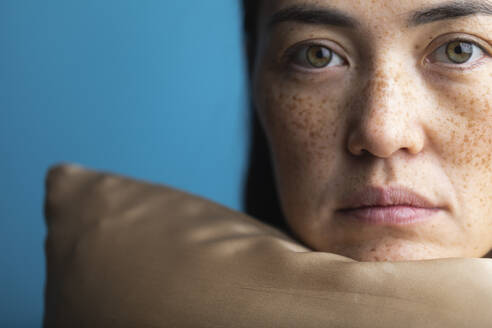 Woman with freckles resting chin on silk pillow against blue background - MRAF00930
