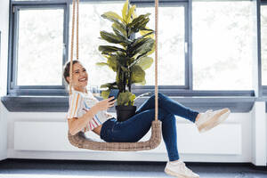 Glückliche Geschäftsfrau mit Mobiltelefon und Topfpflanze auf einer Schaukel sitzend - JOSEF17415