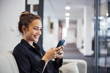 Happy businesswoman using smart phone on sofa in office - JOSEF17442