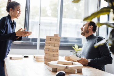 Smiling businesswoman with colleague stacking up wooden blocks in office - JOSEF17448