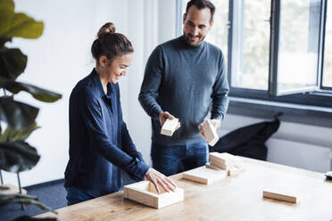 Happy businesswoman with colleague arranging wooden blocks on desk - JOSEF17449