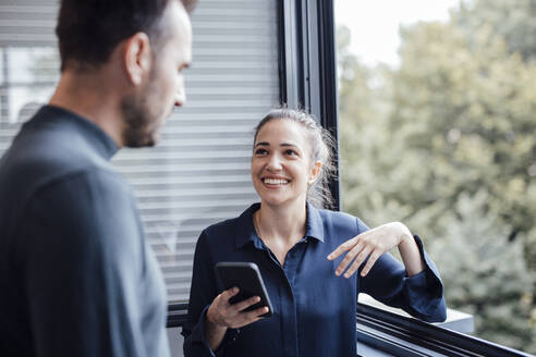Lächelnde Geschäftsfrau steht mit einem Kollegen am Fenster im Büro - JOSEF17466