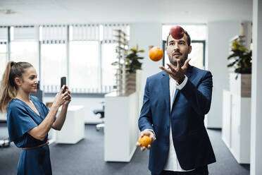 Young businesswoman photographing colleague juggling fruits through smart phone in office - JOSEF17514